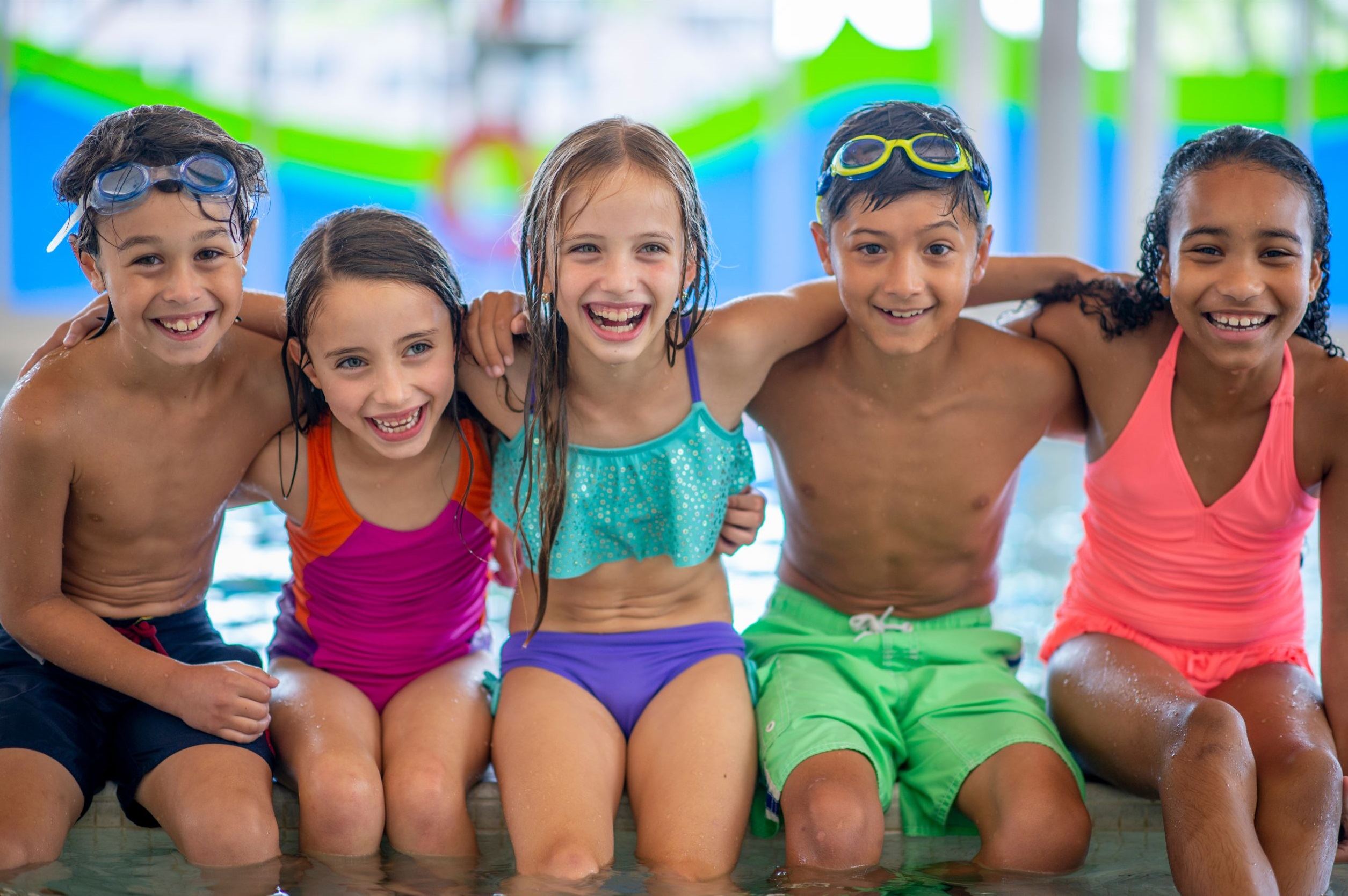 Five children in swim suits interlock their arms while sitting on the edge of a pool 